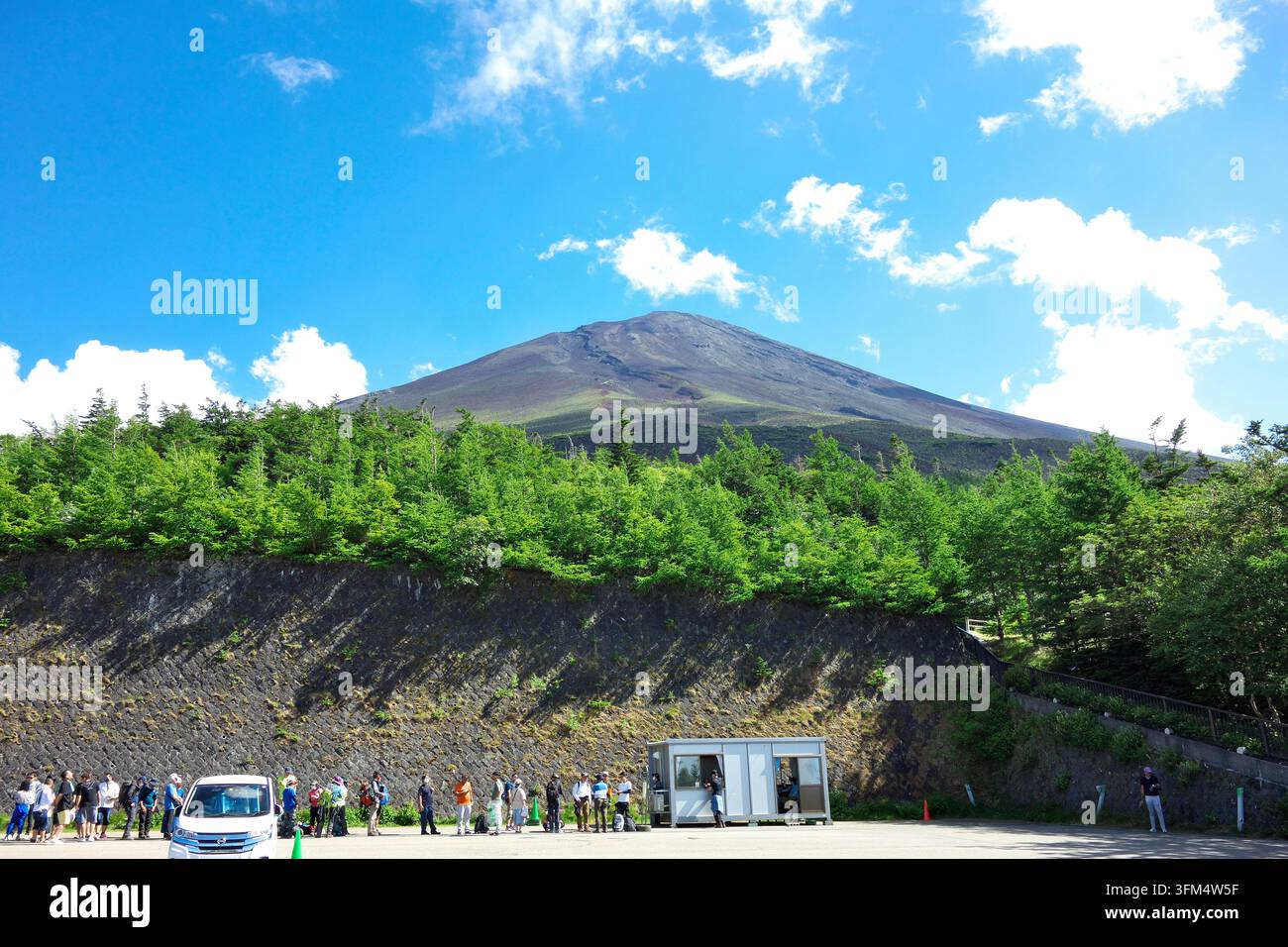 Una vista panoramica del Monte Fuji sotto un cielo blu brillante con nuvole bianche, con turisti e visitatori riuniti in un parcheggio vicino ai sentieri montani Foto Stock