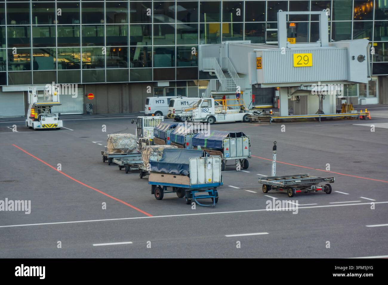 Rimorchi per bagagli dell'aeroporto e carichi imballati attendono vicino a un ponte di imbarco passeggeri e furgoni di servizio, supportando il trasferimento dei bagagli e la gestione del carico su Foto Stock