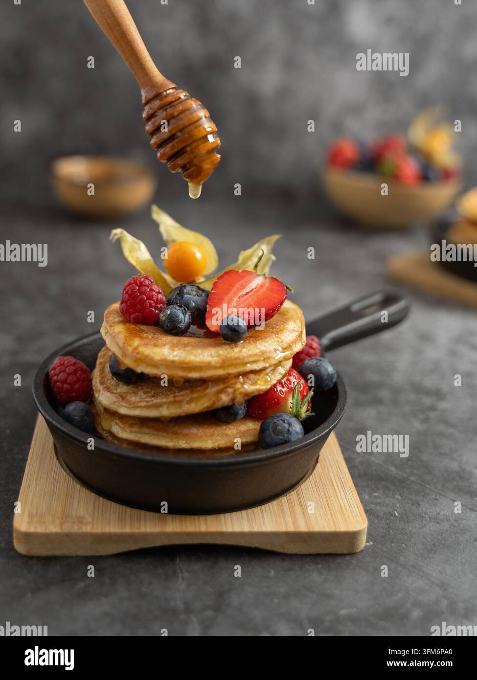 Pila di piccole frittelle ricoperte di frutti di bosco misti e sciroppo di miele, servita in una ciotola scura su una tavola di legno con frutti di bosco aggiuntivi sullo sfondo Foto Stock