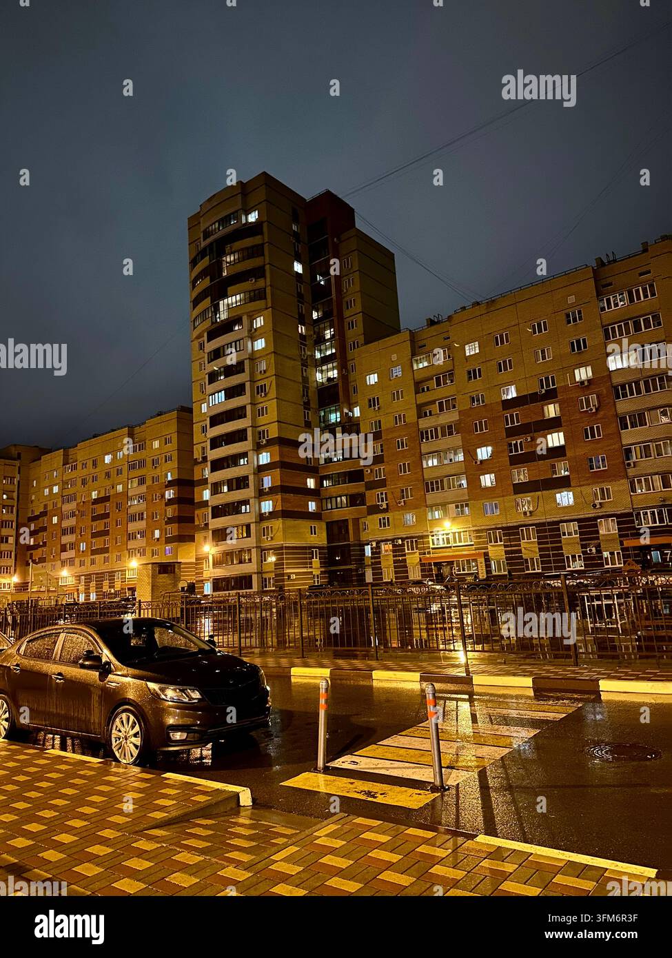 Gli edifici residenziali illuminati si riflettono sulla strada bagnata durante una notte urbana piovosa. - Immagine stock catturata con smartphone
