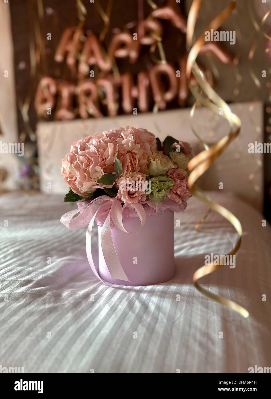 Una scatola di fiori rosa poggia su un letto davanti a decorazioni di compleanno sfocate. - Immagine stock catturata con smartphone