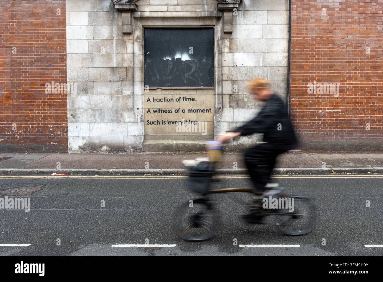 Ciclista sfocato che passa davanti a un vecchio muro di fabbrica con graffiti poetici a Hackney, Londra, Regno Unito Foto Stock