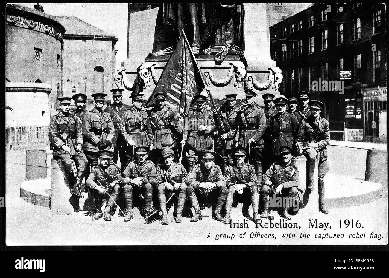 Ufficiali dell'esercito britannico si riuniscono davanti al Monumento di Parnell su Sackville Street, a Dublino, nel maggio 1916 dopo la repressione della Rivolta di Pasqua. I soldati in uniforme posano orgogliosamente attorno a una bandiera ribelle irlandese catturata con una arpa dorata, esibita come trofeo di guerra dopo intensi combattimenti nelle strade della capitale. Questa cartolina souvenir di propaganda esemplifica il trionfalismo imperiale britannico immediatamente dopo l'insurrezione, documentando il dominio militare e la sconfitta dei ribelli in Irlanda. Foto Stock