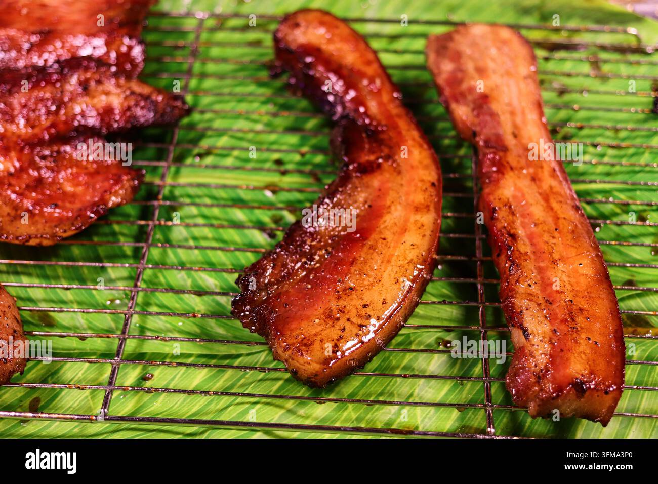 Lunghe strisce di pancetta di maiale grigliata su una griglia metallica sopra un vassoio con stampa a foglie verdi in Thailandia, primo piano, orizzontale, senza persone Foto Stock