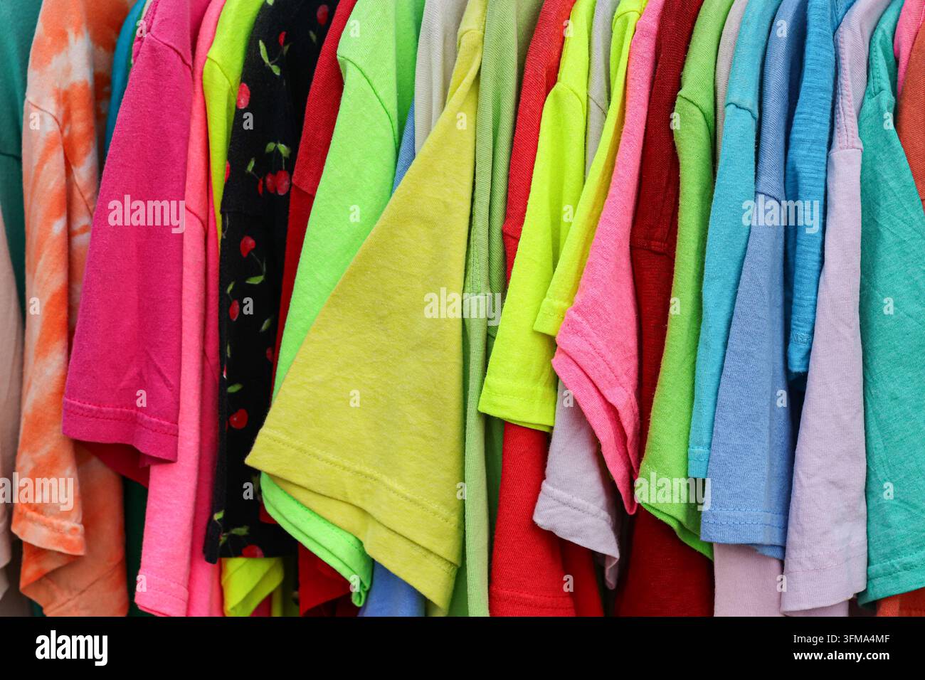 Magliette di seconda mano colorate appese una accanto all'altra in un negozio di seconda mano in un mercato all'aperto in Thailandia, primo piano a campo pieno. Foto Stock