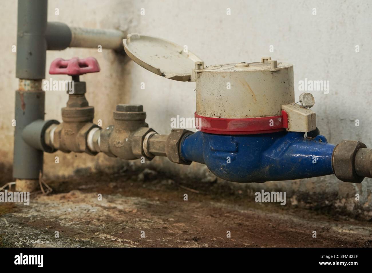Primo piano di un contatore dell'acqua esterno collegato a tubi con una valvola a maniglia rossa. Il sistema sembra funzionante ma mostra segni di età e intemperie. Foto Stock