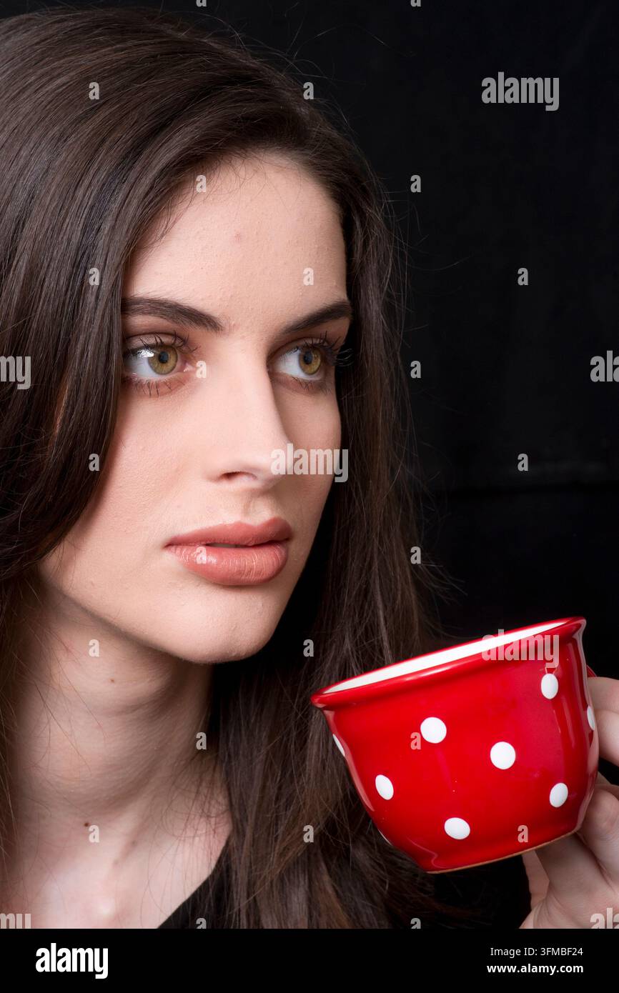 Una giovane donna pensierosa tiene una tazza a pois rossi vicino alle labbra, guardando lontano in un ritratto tranquillo e contemplativo che trasmette calma, introspezione e Foto Stock