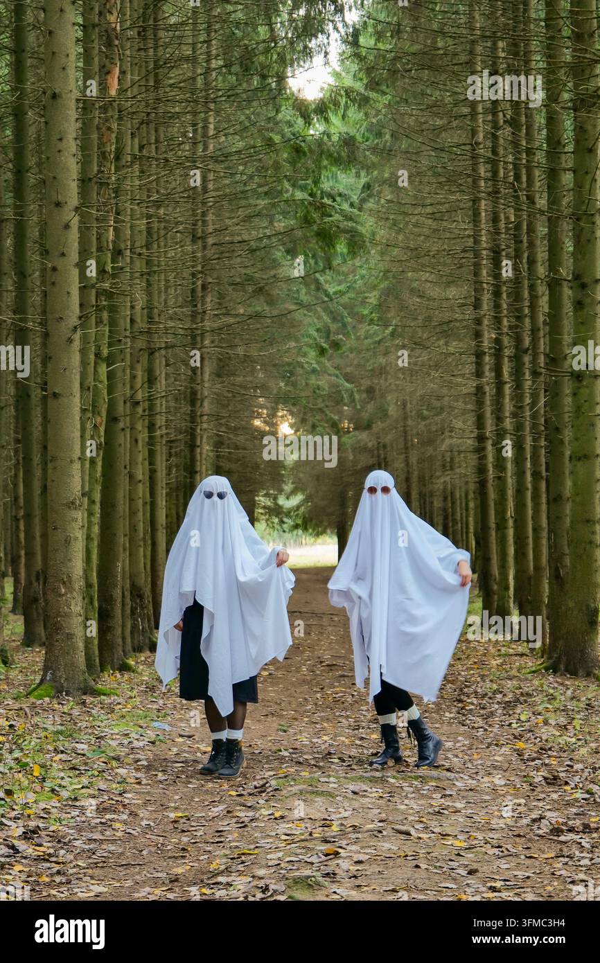 Due adulti in costumi da fantasmi bianchi posano su un sentiero nel bosco, gli occhiali da sole sono visibili attraverso le coperture di lenzuolo. Coppia giocosa di Halloween indossa abiti scuri e un "buu"} Foto Stock