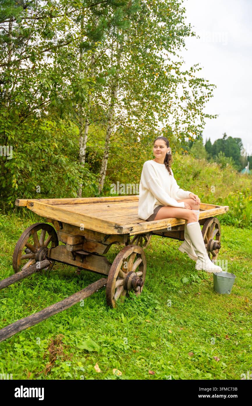 Giovane donna in maglione bianco e calzini al ginocchio seduta su un carretto di legno in un campo verdeggiante circondato da alberi e cielo nuvoloso Foto Stock