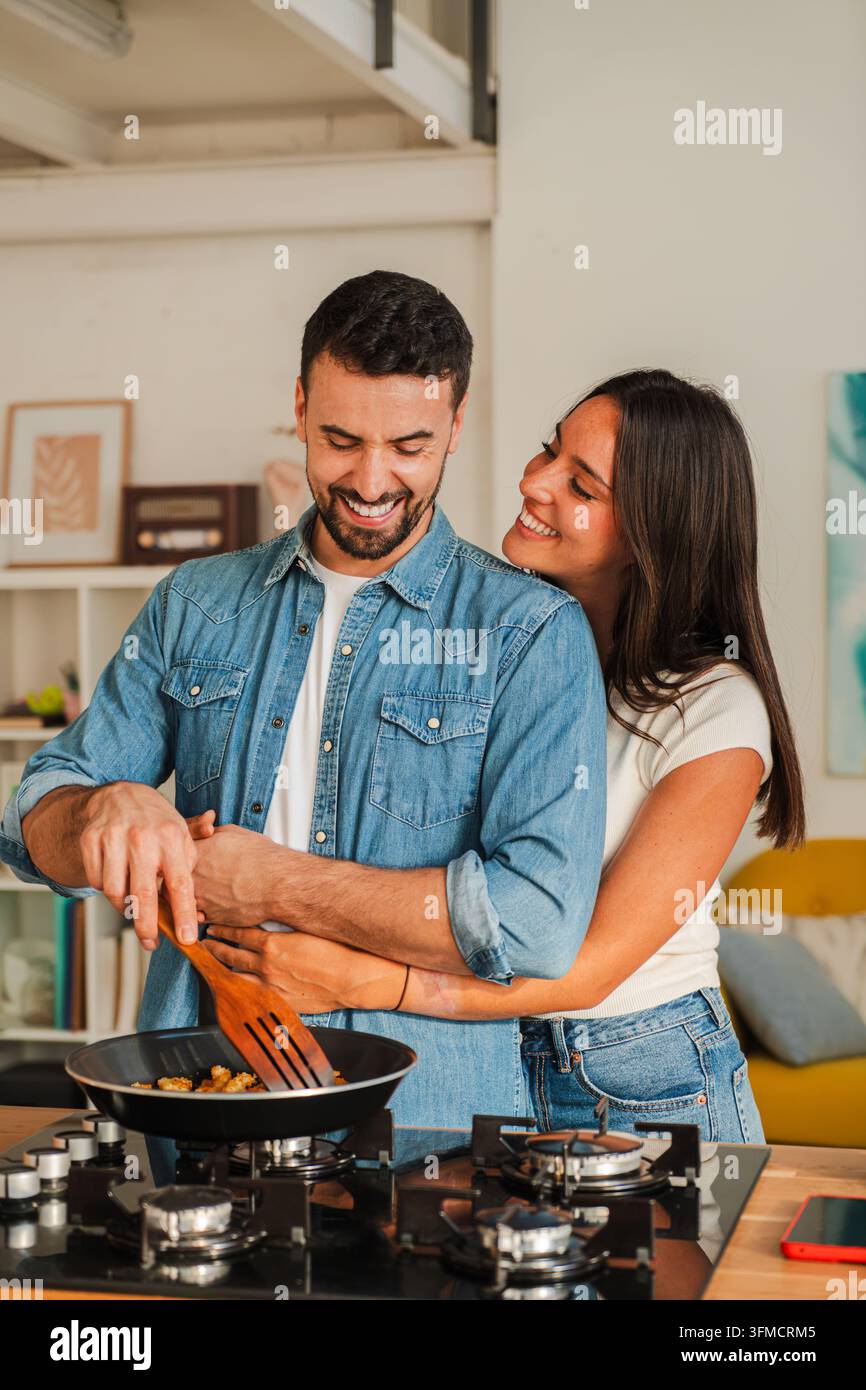 Due persone ridono cucinando insieme su un fornello compatto, uno mescola verdure con una spatola di legno, la macchina fotografica cattura mani allegre e una casa calda Foto Stock