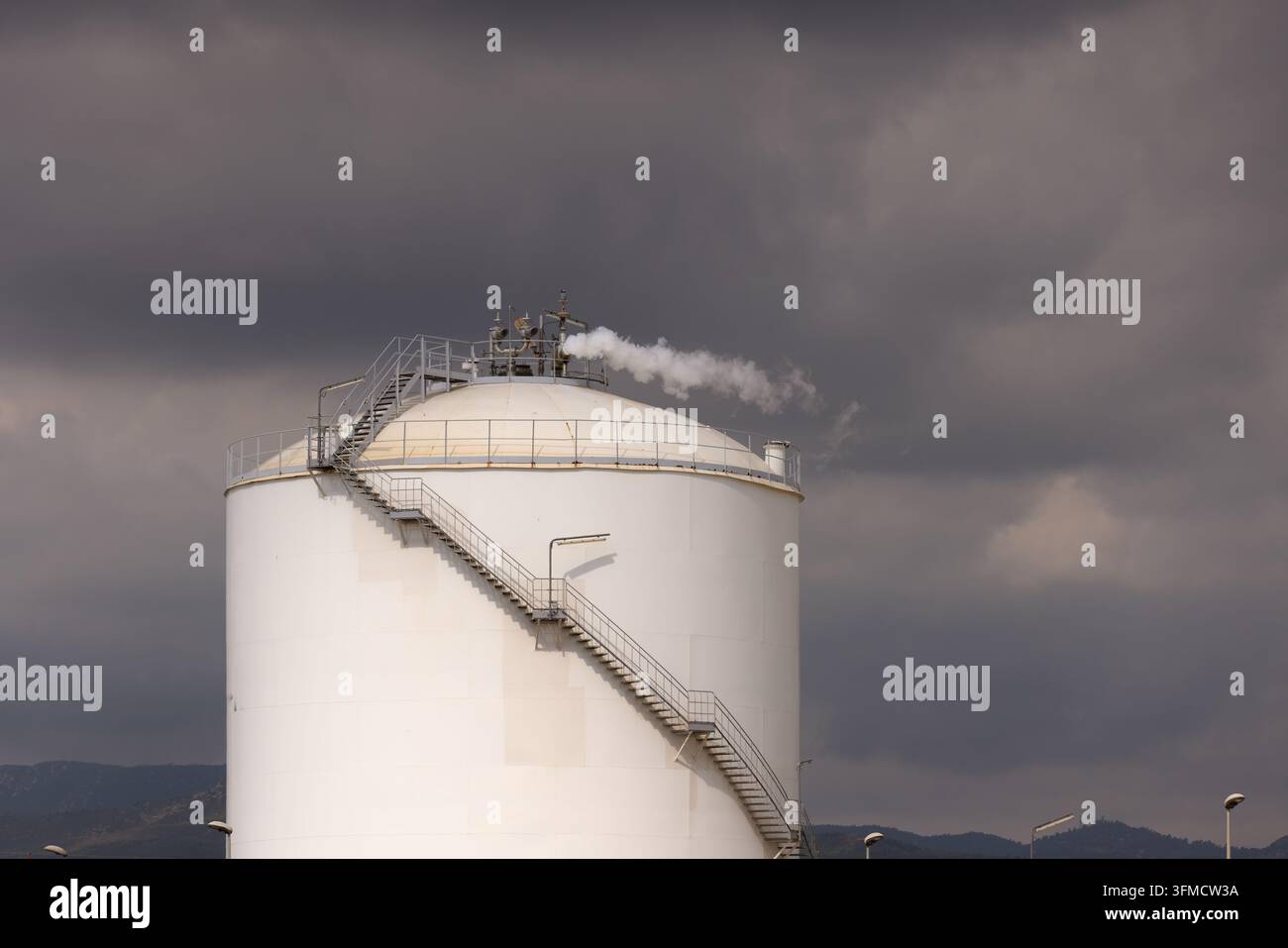 Grande serbatoio cilindrico di olio bianco con scala a spirale in metallo che rilascia vapore contro un drammatico cielo nuvoloso scuro. Foto Stock