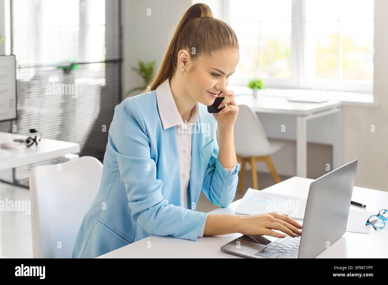 Giovane analista che parla al telefono mentre digita su un laptop alla scrivania dell'ufficio Foto Stock