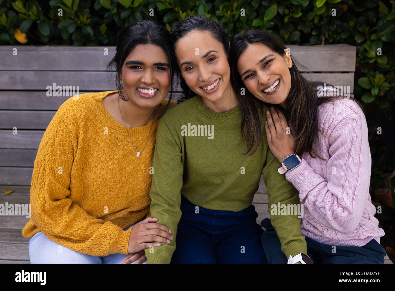 Diverse amiche sedute su una panchina a doghe nel parco, indossano maglioni di lana e un ciondolo smartwatch Foto Stock