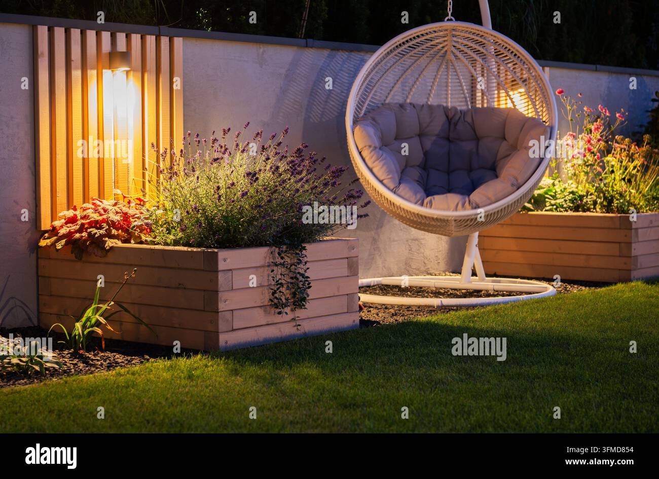 Una sedia sospesa imbottita a forma di uovo si trova accanto a vasi di fiori in legno e illuminazione calda del giardino di notte. Foto Stock