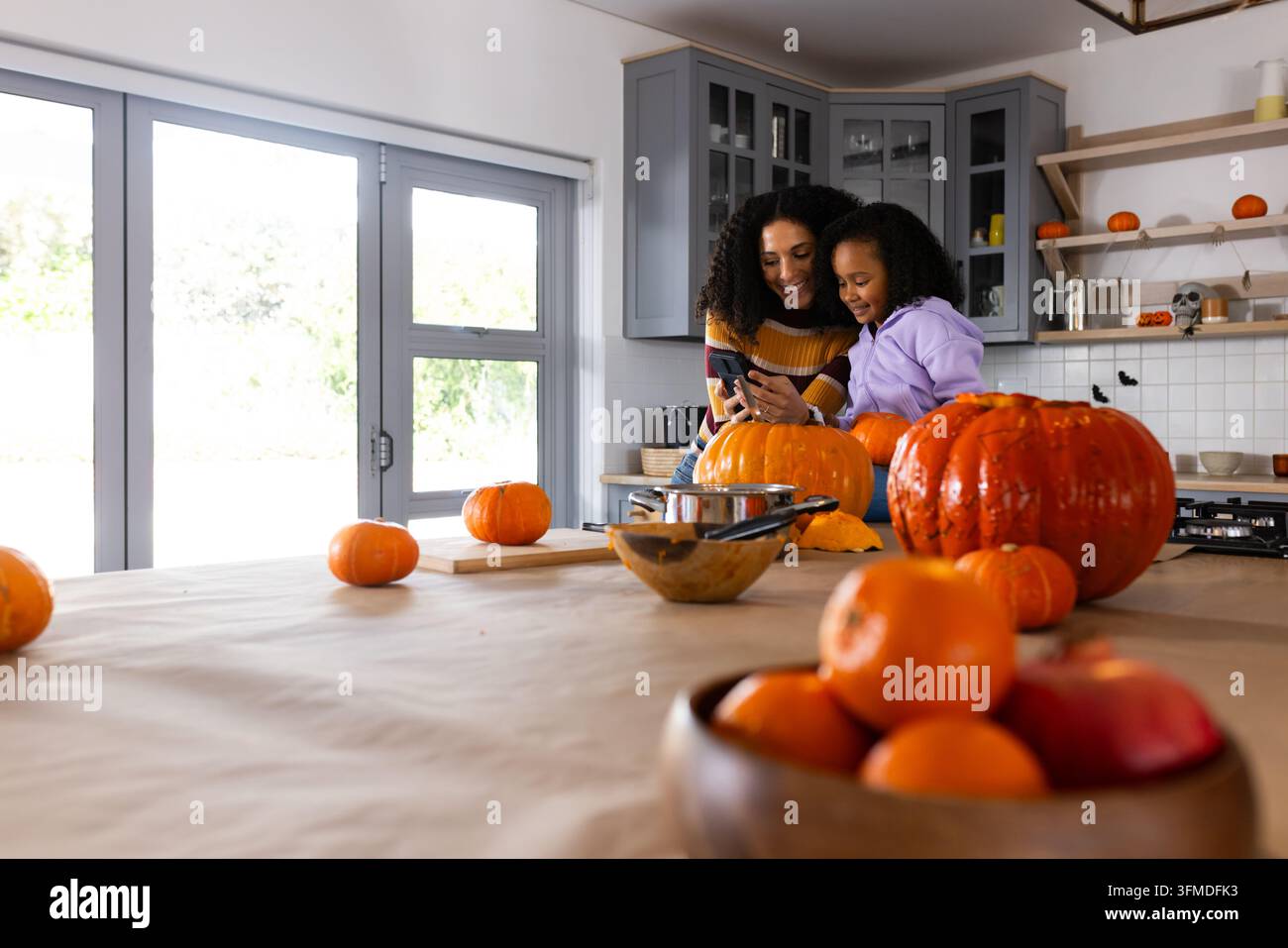Mamma afroamericana e figlia adolescente che intagliano una zucca di Halloween usando il cellulare in cucina, spazio di copia Foto Stock
