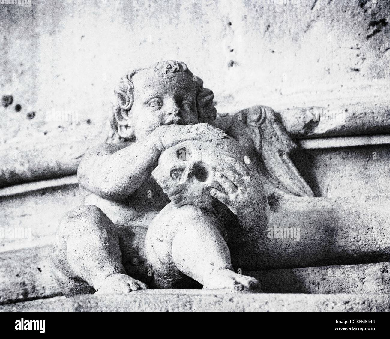 Foto in bianco e nero di una scultura in pietra di un cherubino che tiene un cranio umano, raffigurante un classico motivo di memento mori su una facciata architettonica antica Foto Stock