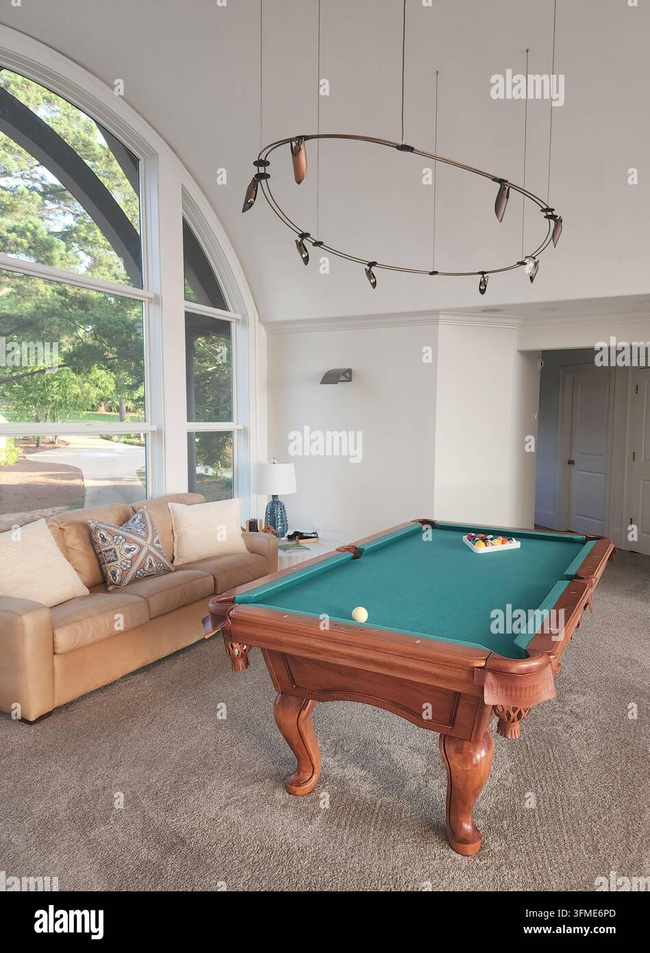 Ampio interno di una sala giochi di lusso con un classico tavolo da biliardo in legno, divano in pelle e moderno lampadario ovale vicino a una grande finestra ad arco Foto Stock