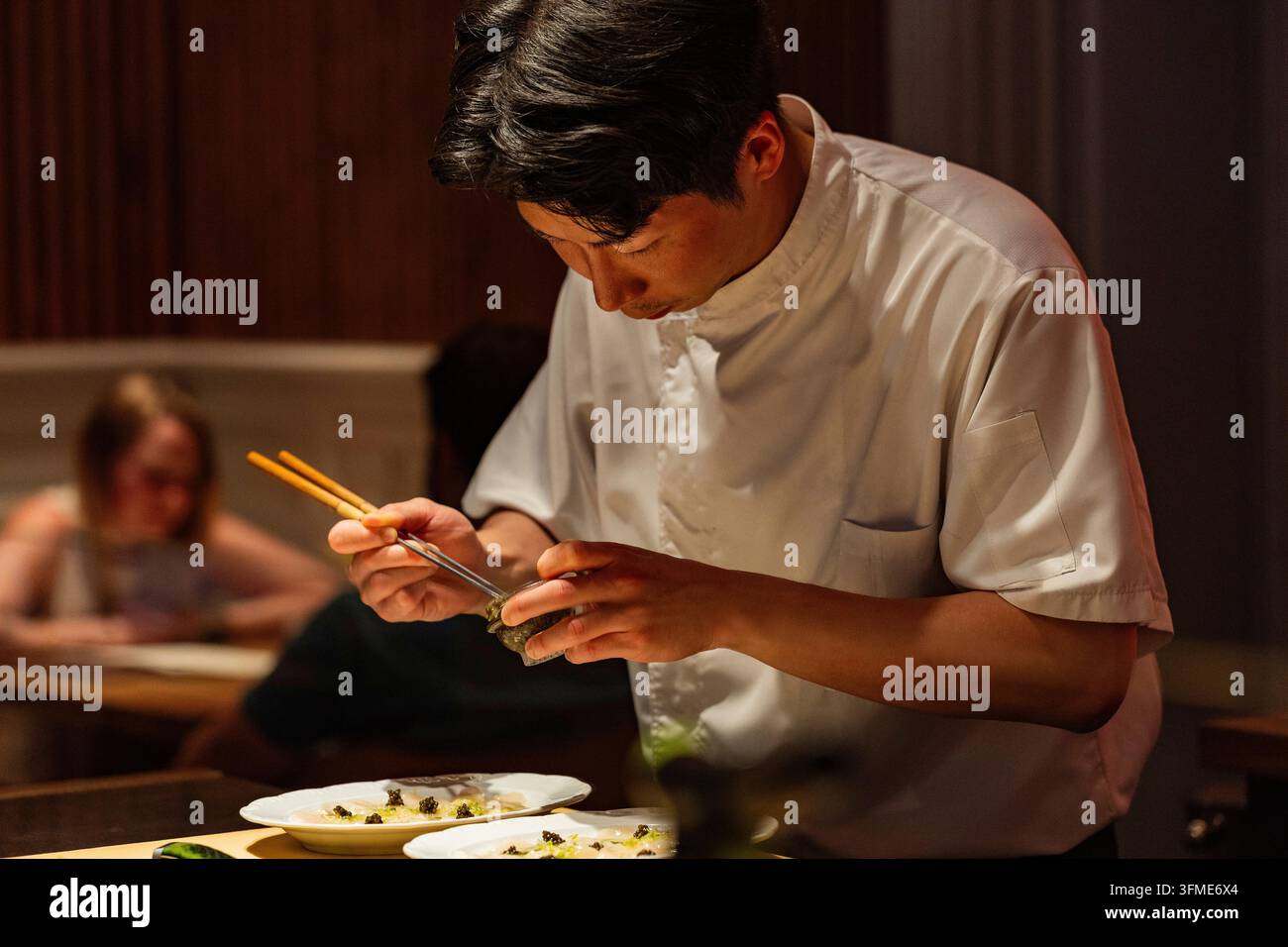 Berlino, Germania – 19 agosto 2025: Uno chef maschile asiatico concentrato prepara abilmente piatti di sushi con le bacchette in un ristorante scarsamente illuminato, mostrando Foto Stock