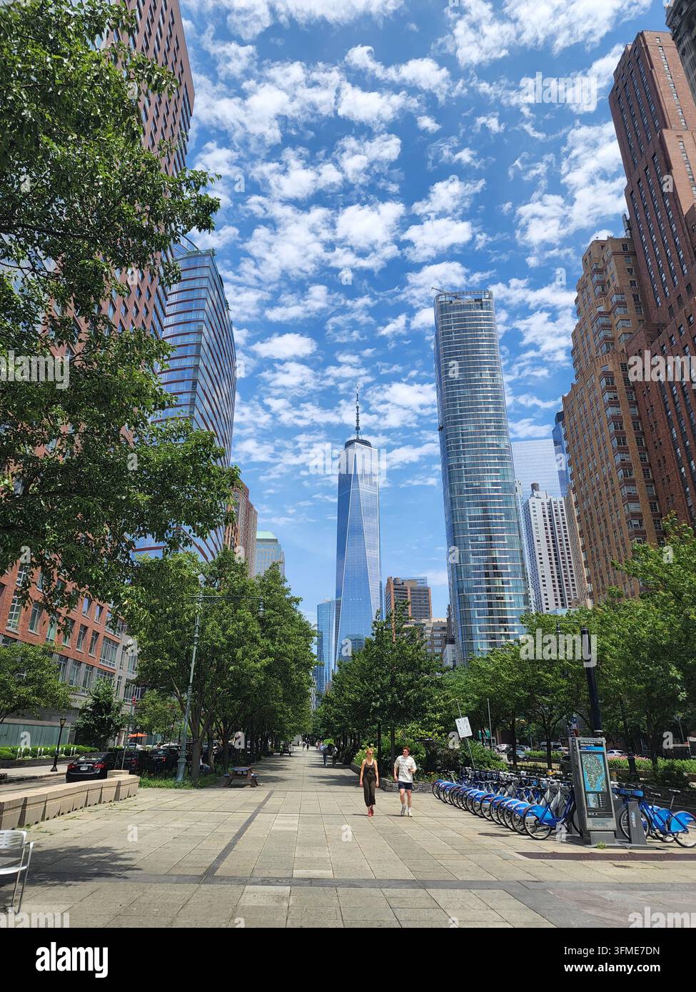NEW YORK CITY – 26 GIUGNO 2024: Pedoni camminano lungo un viale pedonale verde a Battery Park City con il One World Trade Center sullo sfondo. - Immagine stock catturata con smartphone