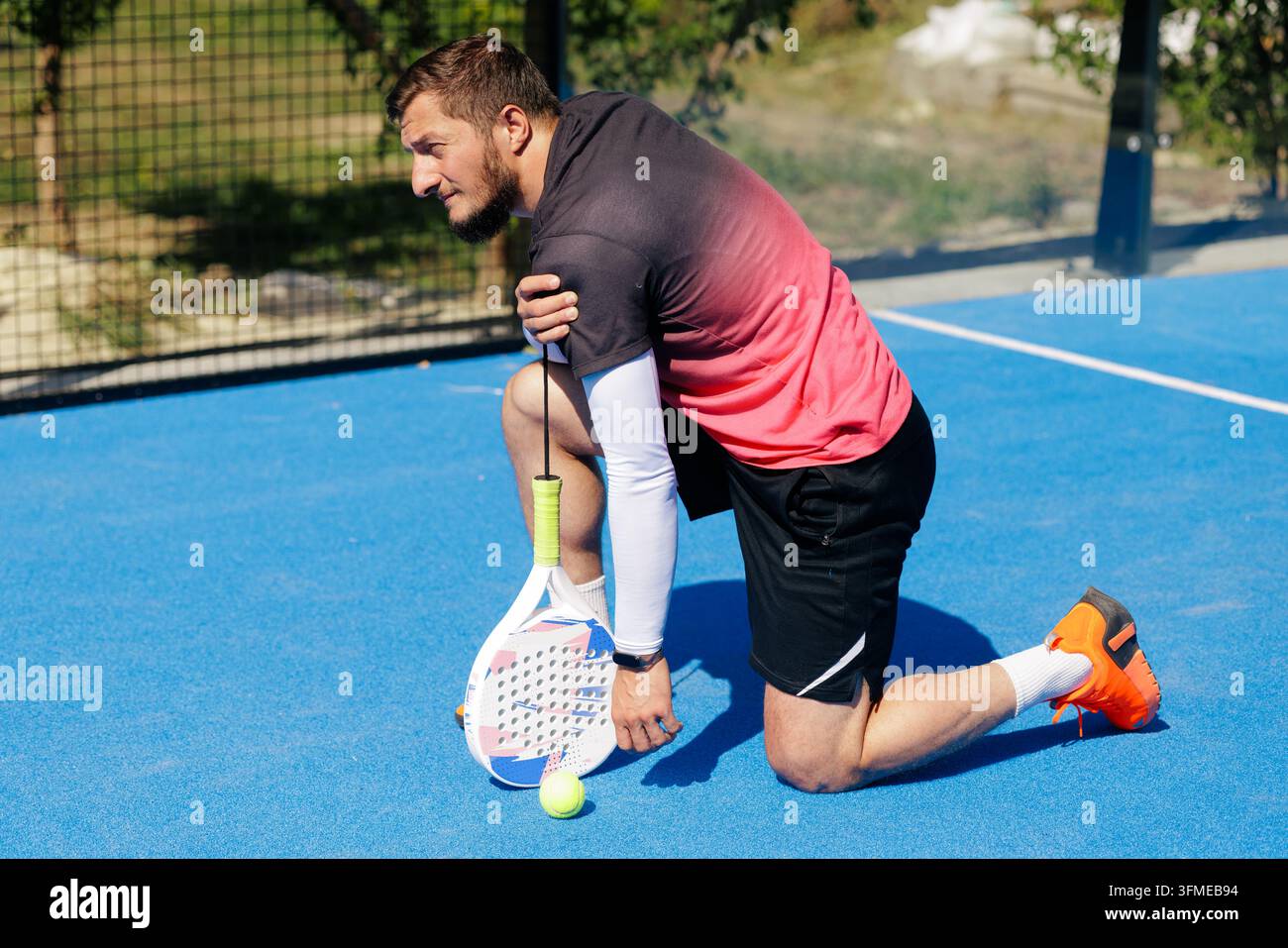 Giocatore di padel infortunato inginocchiato su un campo all'aperto mentre tiene la spalla, illustrando il dolore sportivo, lo sforzo fisico e la prevenzione degli infortuni per la salute Foto Stock