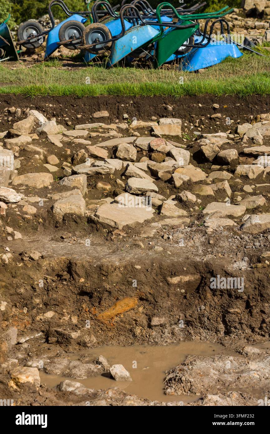 Trincea di scavo che mostra lastricioli di pietra esposti e strati di terreno sovrapposti, fila di carriole rovesciate sopra il pendio erboso in uno scavo archeologico Foto Stock