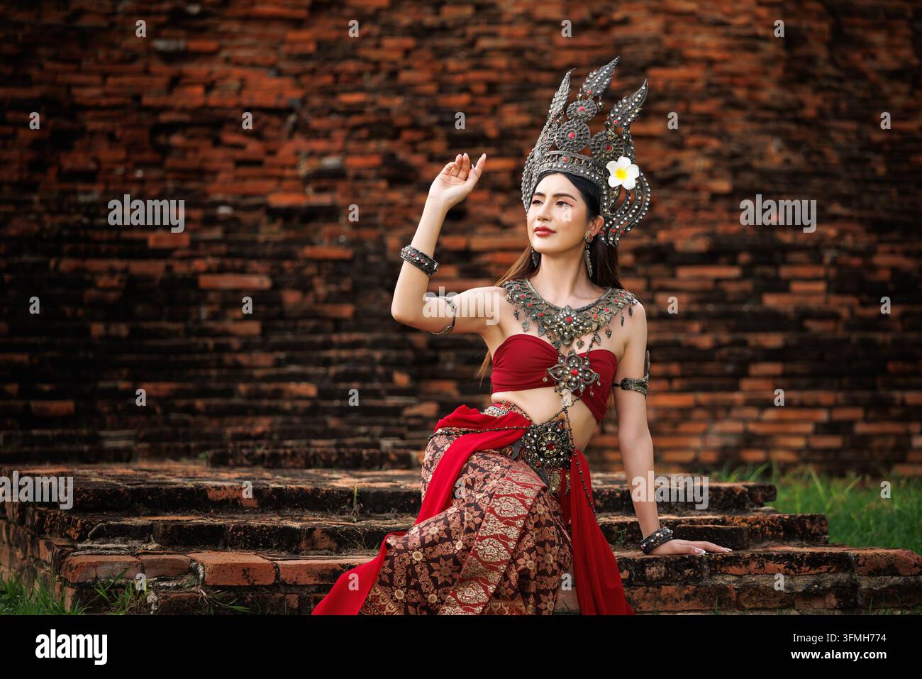 Donna asiatica in autentico costume tradizionale e copricapo ornato seduta all'aperto presso rovine storiche in mattoni. Foto Stock