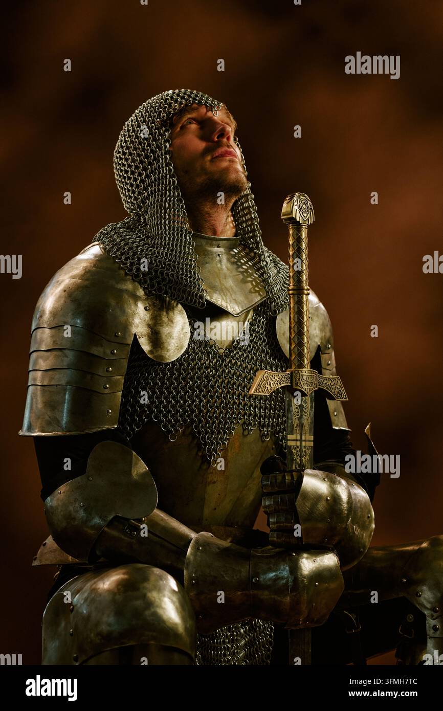Cavaliere medievale in armatura inginocchiato e in preghiera con una spada Foto Stock
