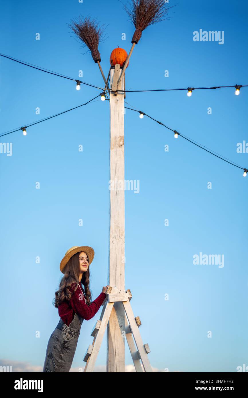 Giovane donna con cappello di paglia accanto a un alto palo di legno con zucca e scope, luci a filo luminose creano una scena festiva di Halloween su uno sfondo blu chiaro Foto Stock