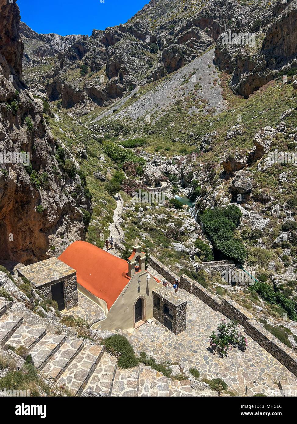 Vista ad angolazione alta di una piccola cappella con tetto arancione e scalini in pietra nella gola rocciosa di Kotsifou, Creta, Grecia, sotto un cielo blu brillante. - Immagine stock catturata con smartphone