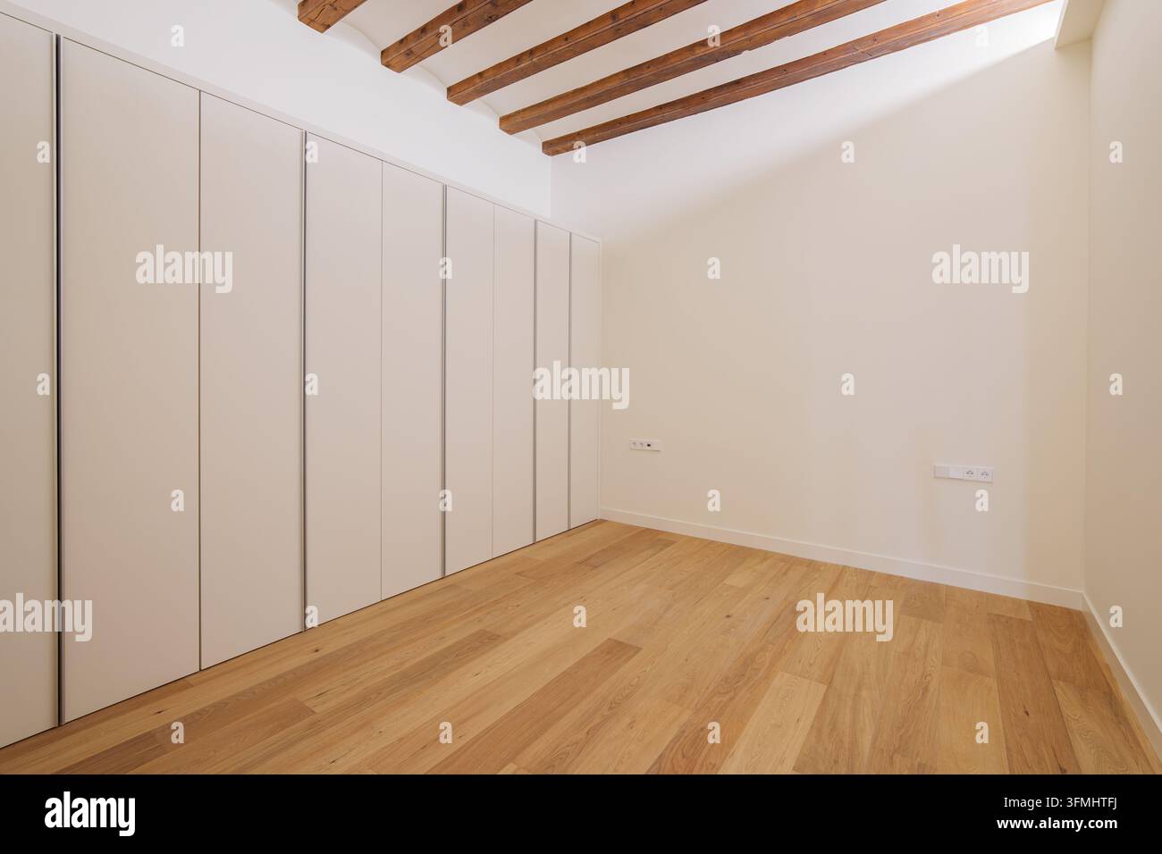 Stanza minimalista vuota con pavimento in quercia chiara, pareti bianche, travi a vista sul soffitto e armadi built-in a tutta altezza con ante a filo Foto Stock
