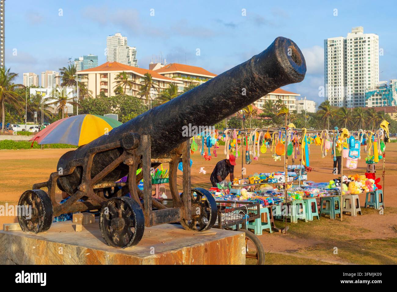 Sri Lanka, Colombo, Galle Face Green: un cannone dell'era coloniale con una bancarella di strada che vende giocattoli e edifici sul lungomare sullo sfondo Foto Stock