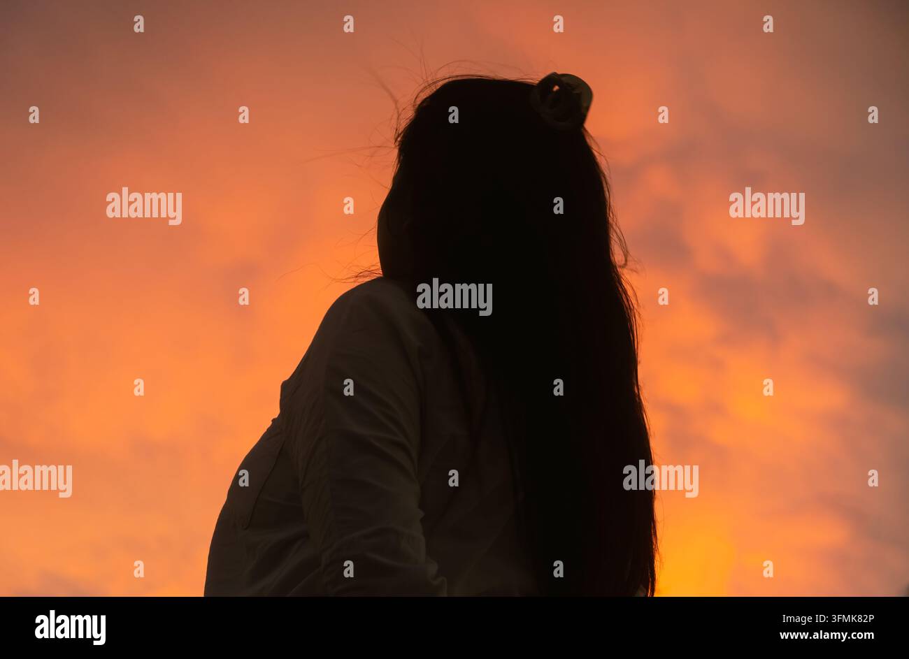 Silhouette di una donna asiatica con lunghi capelli che guarda un cielo arancione vibrante al tramonto Foto Stock