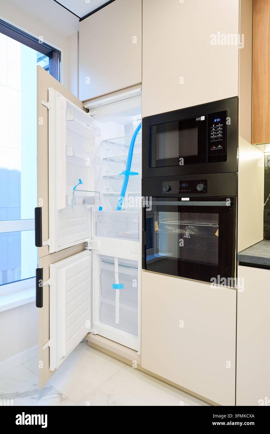 Frigo aperto integrato accanto a forno e torre microonde in un interno di cucina moderno. Design di appartamento elegante con elettrodomestici integrati, mobili minimalisti e concetto di archiviazione premium Foto Stock