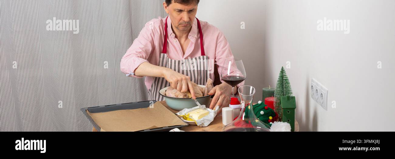 Un uomo di 53 anni sta preparando il pollo per la cena pre-natalizia. Condisce il pollo in una ciotola. Nelle vicinanze ci sono patate e un bicchiere di vino sul tavolo Foto Stock
