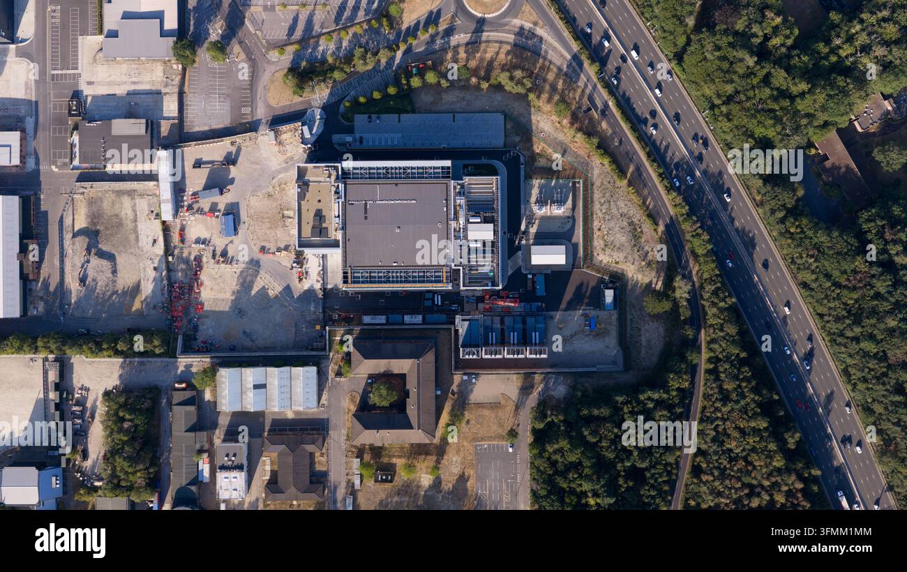 Vista aerea di una grande struttura industriale con tetto piatto, aree di parcheggio circostanti e zone di costruzione, situata adiacente a una superstrada a più corsie Foto Stock