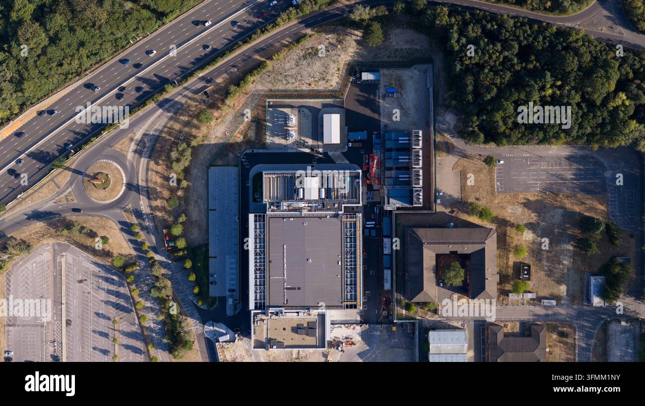 Vista aerea di una moderna struttura industriale con un grande edificio a tetto piatto, aree di parcheggio adiacenti e un rondò autostradale nelle vicinanze a Longcro Foto Stock