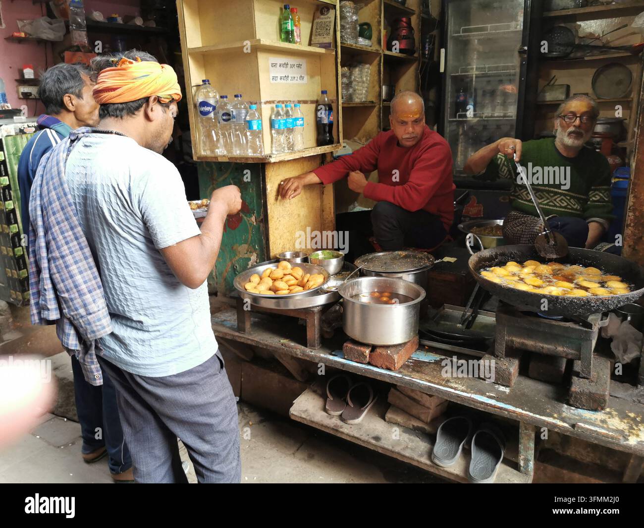 VARANASI, INDIA - 22 GENNAIO 2026: I venditori frigono i kachori in una grande padella a uno stand di street food in un vicolo mentre un cliente tiene un piatto Foto Stock