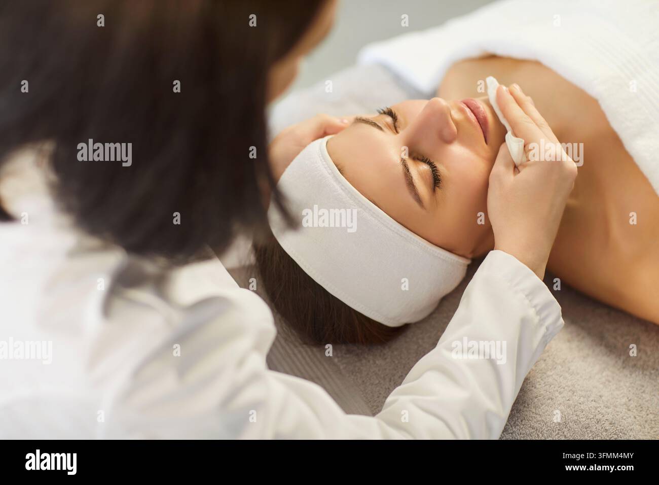 Giovane donna estetista che pulisce il viso di una donna con un tovagliolo, idratando la pelle Foto Stock