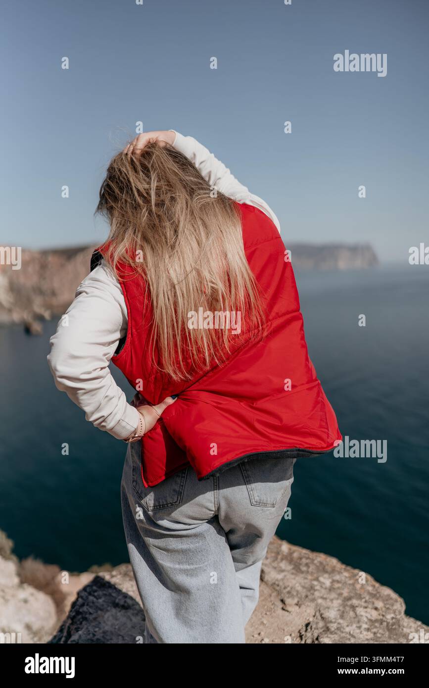 Punto di vista sull'oceano da scogliera di un viaggiatore cattura una donna bionda in un giubbotto rosso in piedi su un bordo roccioso che guarda il mare Foto Stock