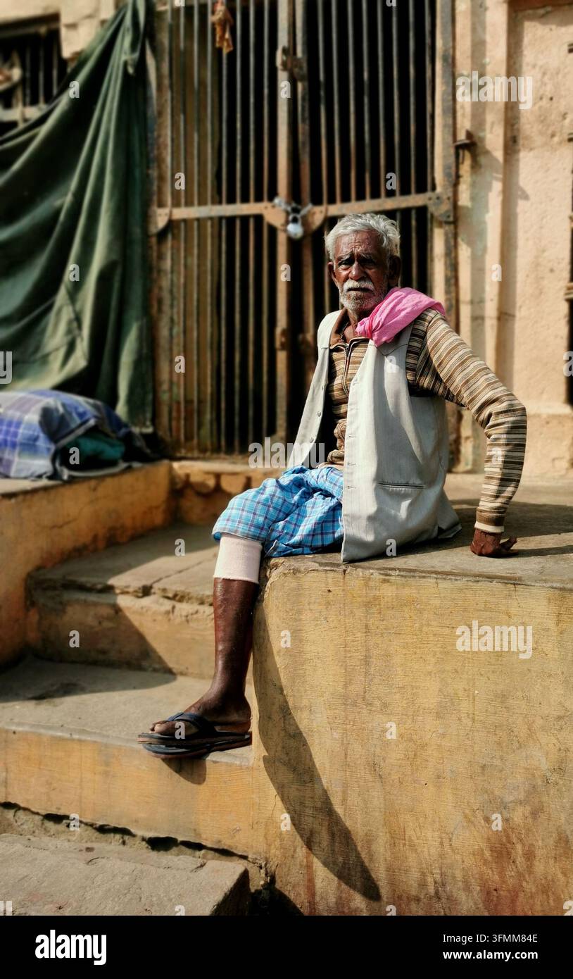 VARANASI, INDIA - 22 GENNAIO 2026: Un uomo anziano in maglione a righe, gilet e lungi a quadri è seduto su un muretto di pietra accanto a una scalinata Foto Stock