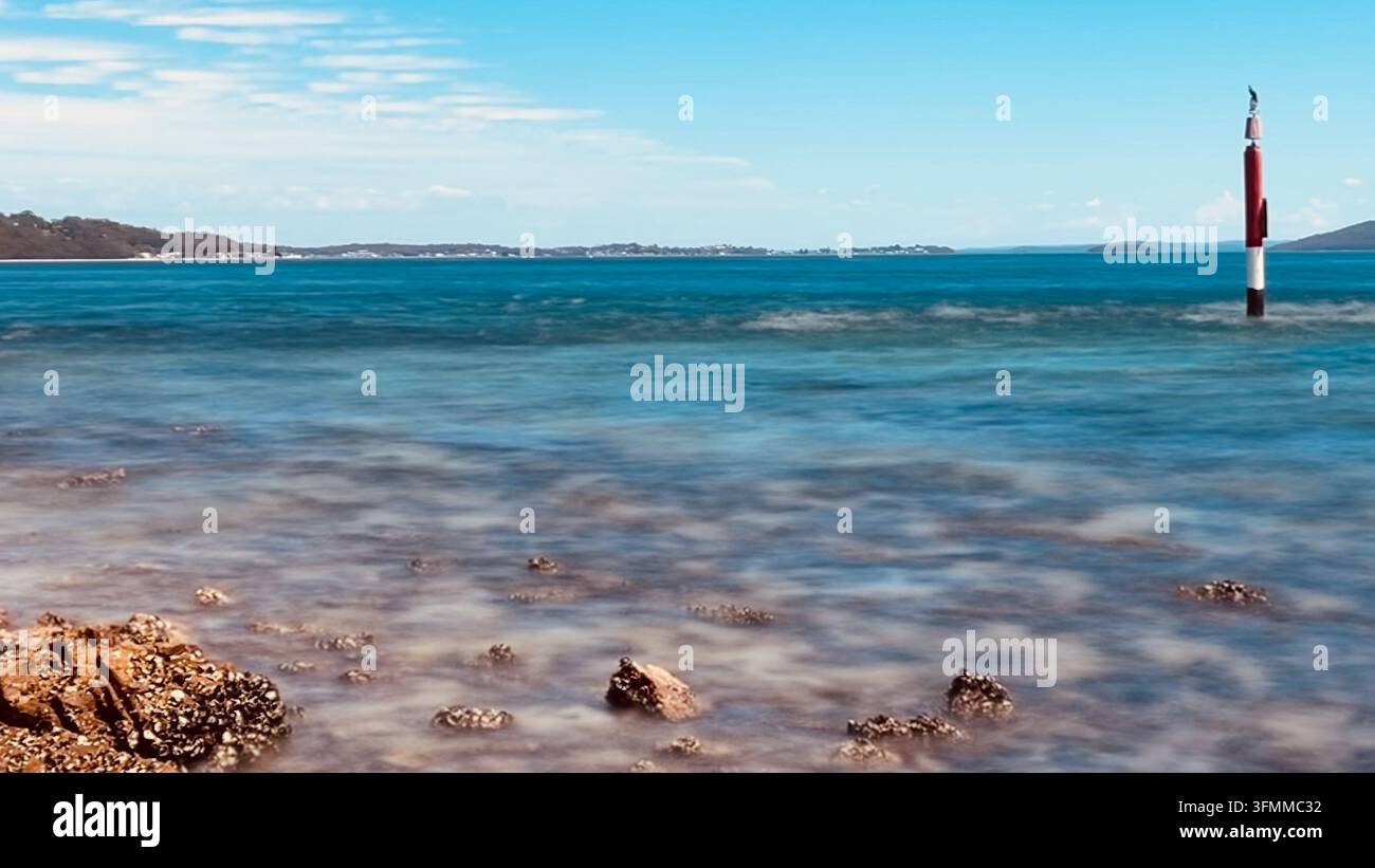 L'esposizione lunga ammorbidisce l'acqua turchese intorno a una formazione rocciosa, un segnalatore di navigazione a strisce che si erge nella baia di Port Stephens, NSW, Australia Foto Stock