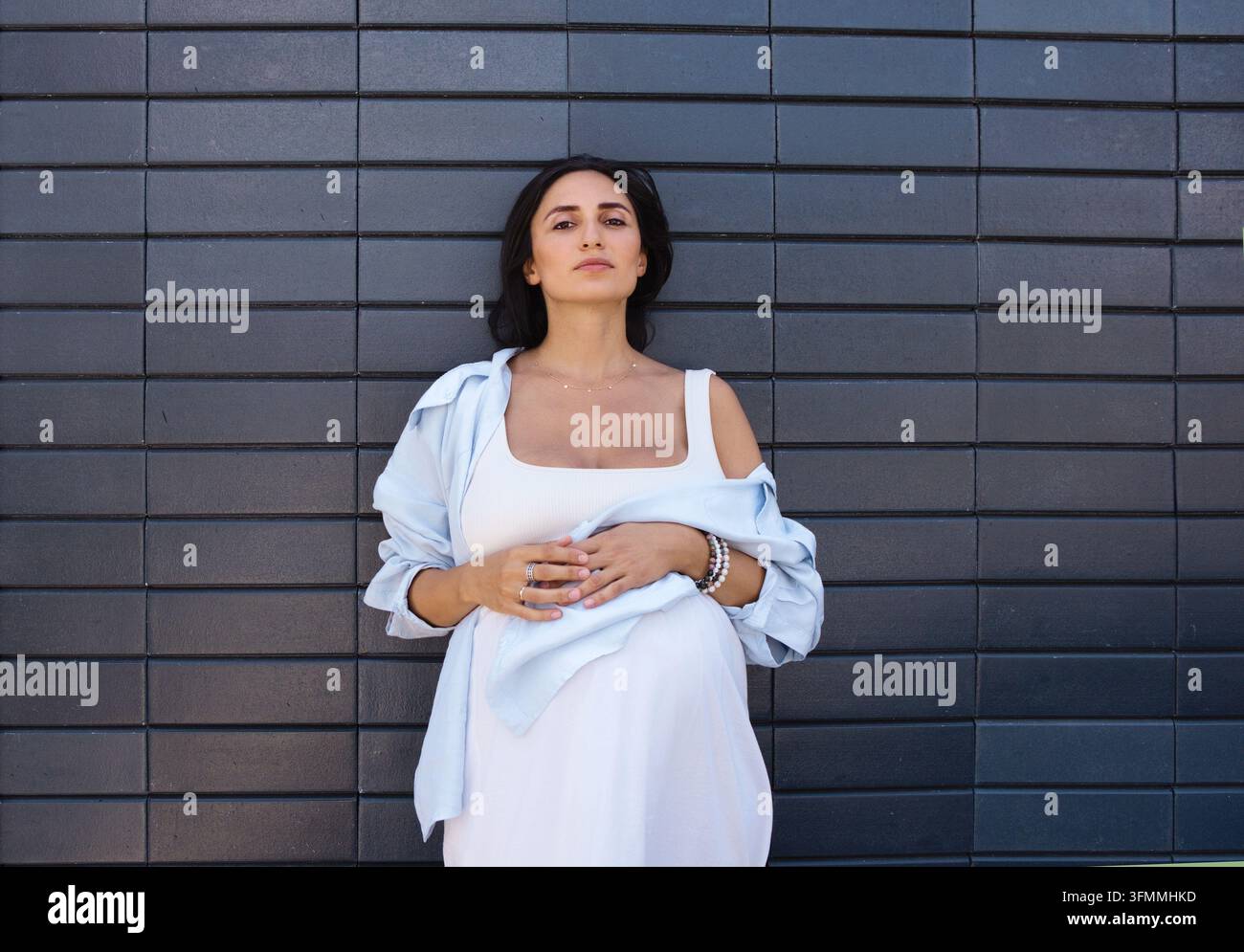 Donna incinta in abiti bianchi appoggia le mani sul ventre mentre è in piedi al centro davanti a un muro esterno orizzontale piastrellato scuro Foto Stock
