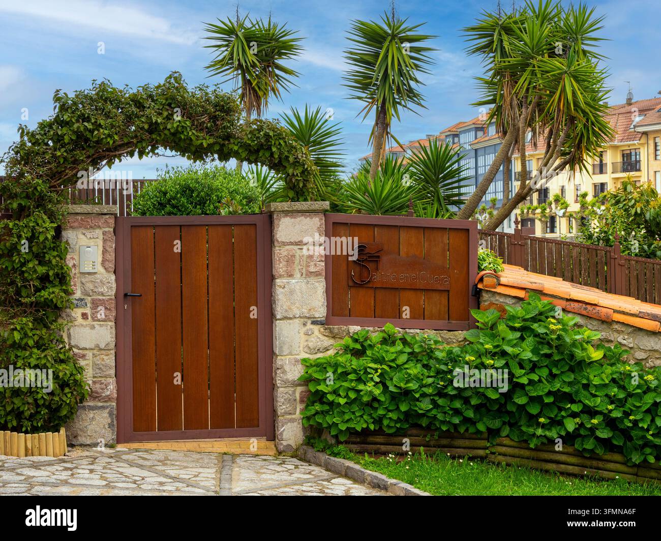 Cortile della casa di pietra con cancello in legno - Esterno elegante di una casa di pietra con cortile lastricato, cancello da giardino in legno, vegetazione rigogliosa e palme sotto il blu Foto Stock