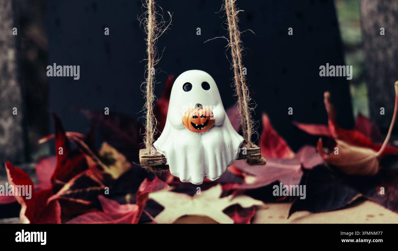 Decorazione Halloween carina con un fantasma giocattolo che tiene un zucca su un'altalena con foglie rosse sotto. Ottima per il marketing stagionale e il blogging delle feste. Foto Stock