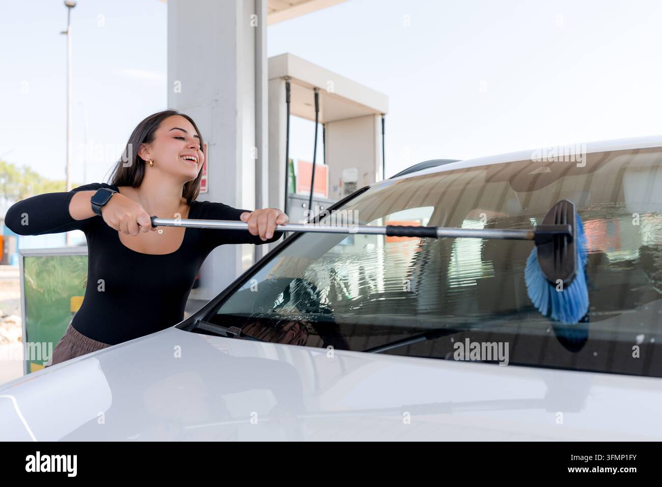 Donna sorridente usa una spazzola a manico lungo per pulire il parabrezza di un'auto bianca in una stazione di lavaggio self-service, mantenendo il suo veicolo all'aperto. Foto Stock