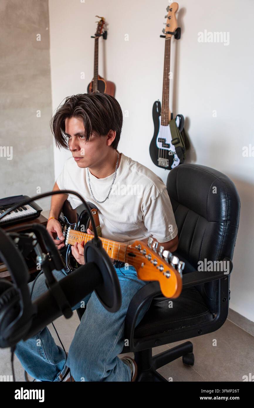 Giovane compositore di musica suona una chitarra elettrica a una scrivania in uno studio domestico, circondato da strumenti, attrezzature per tastiere e microfono per la creatività Foto Stock