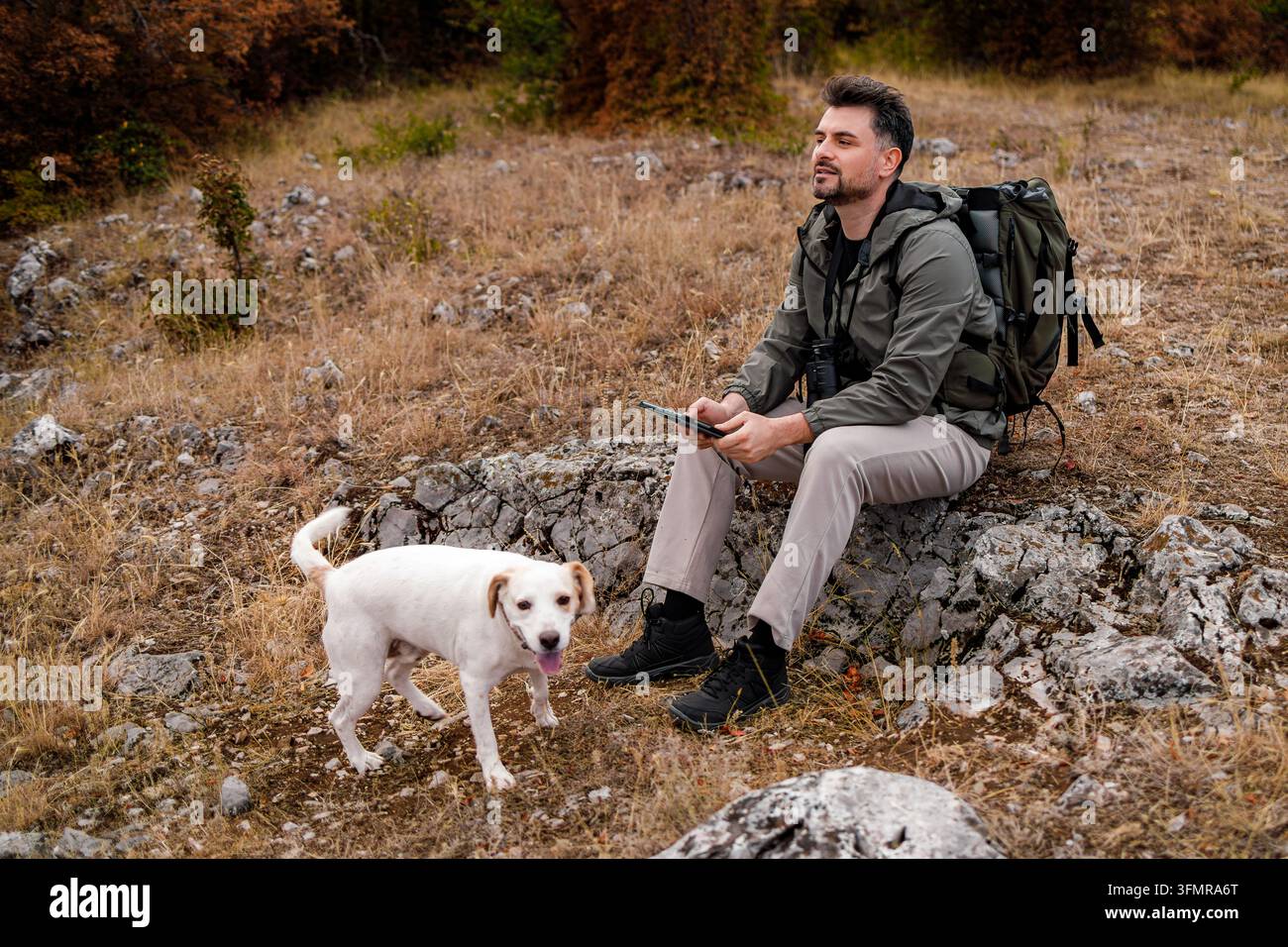 Un uomo è seduto su una roccia e guarda un dispositivo con un cane nelle vicinanze in un campo erboso. Foto Stock