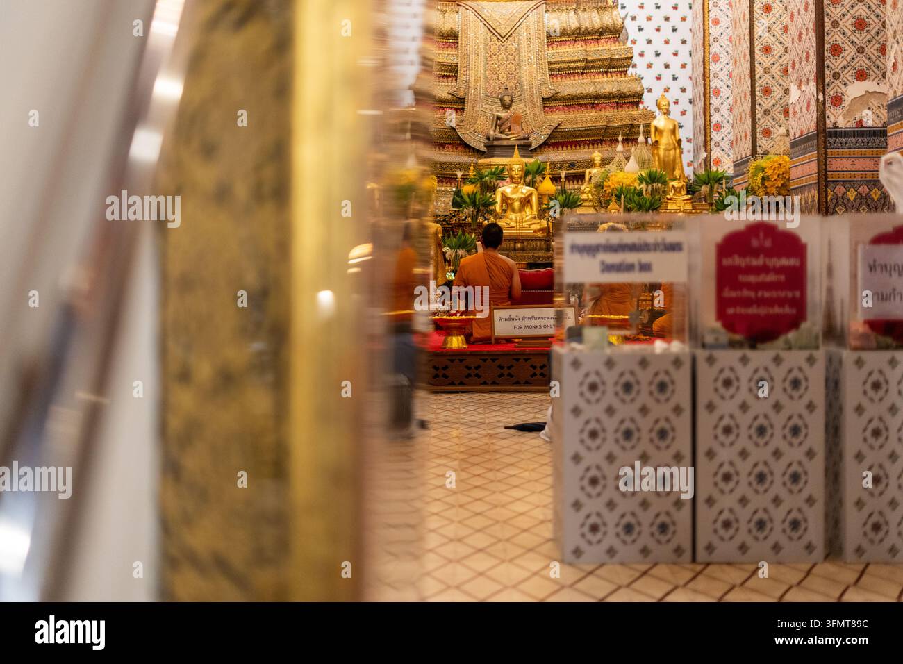 Monaco in abiti arancioni seduto in un tempio thailandese, circondato da statue di Buddha dorate e offerte Foto Stock