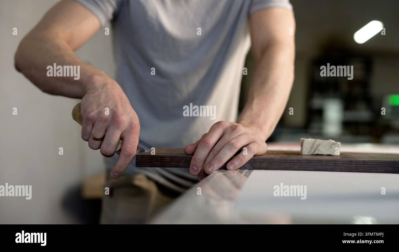 Mani di falegname che rifiniscono con cura il bordo di una tavola di legno scuro con uno strumento manuale in officina, lavoro manuale esperto Foto Stock