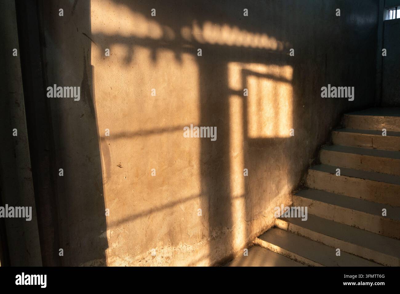 Un angolo interno scarsamente illuminato con una scala ascendente e un raggio di sole che crea modelli di ombra drammatici sul muro. Foto Stock